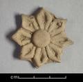Fragment of wall-decoration made of yellowish grey clay with traces of red pigments visible. The rosette-shaped ornament has a raised boss and eight pointed petals. The centre of each petal is incised with a deep line.