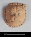 Round clay figure depicting a lion's face. The animal is shown in full frontal view with its head surrounded by the mane. The representation is very stylised. The fact that the figure is flat on the reverse side suggests that it was applied to another object, probably a vessel.;