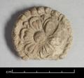 Stucco ornament in the shape of a six-petalled lotus with a raised seed cup, set within a bead border. Traces of a white slip and red pigments are clearly visible.