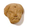 Clay plaque showing the face of a man. He has short hair, a moustache and a concentric circle on his forehead. Details such as eyebrows, moustache and the strands of hair were rendered using incised lines. The object was most likely used as an appliqu, for instance for a vessel.;