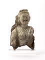 Fragmentary stucco figurine. The remaining part is the bust of a female person with a wrinkled face. She seems to be laughing with wide open mouth and half-closed eyes. Her hair is drawn back loosely under a fillet consisting of plain and beaded bands, the topknot of the hairstyle is broken. She is dressed in a clingin robe which seems to be embroidered partly and wears a large round earring in her right ear. Behind the figure, the remains of a cloak are visible. Traces of a white slip and yellow paint are still visible.;