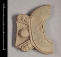 Fragment of wall-decoration made of yellowish brown clay. The remaining part of the ornament is crescent-shaped but the tips are missing. At the base are the remains of a triangle-shaped structure whose sides consist of a row of beads bordered by plain bands. Right in the middle between the two lines sits a large round bead.  The back of the object is plain and smoothed.