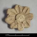 Fragment of wall-decoration made of grey clay with traces of red  pigments visible. The rosette-shaped ornament has a raised boss and eight rounded petals. The centre of each petal is incised with a deep line.