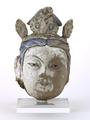 Fragment of a statue made of stucco. This is the head of a Bodhisattva with a curly moustache. The hair is parted from the centre and fastened in a fan-shaped topknot. The Bodhisattva wears an elaborate crown, consisting of a beaded tiara which supports three plaques that are decorated with formalised floral motifs. Traces of pigments make clear that the face was originally painted white, the hair blue and the eyebrows, pupils as well as the moustache black. The crown has red and green details.;