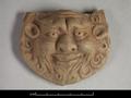 Figure made of clay which as been painted with red colour. Shown here is a grotesque face with a broad nose, bulging eyes and a long beard.