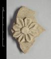 Fragment of wall-decoration made of yellowish grey clay with traces of red  pigments visible. The rosette-shaped ornament has a raised boss and eight petals, partly rounded partly pointed. The centre of each petal is incised with a deep line.