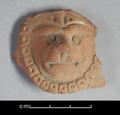 Round clay figure depicting a lion's face. The animal is shown in full frontal view with its head surrounded by the mane. The representation is very stylised. The fact that the figure is flat on the reverse side suggests that it was applied to another object, probably a vessel.;