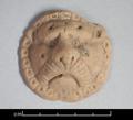 Round clay figure depicting a lion's face. The animal is shown in full frontal view with a moustache and its head surrounded by the mane. The representation is very stylised. The fact that the figure is flat on the reverse side suggests that it was applied to another object, probably a vessel.;