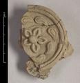 Fragment of statue made of brown clay.The object has the shape of a large round medallion, probably once a decorative ornament of a halo. A flower in the centre is surrounded by a ring of leaves or flames. This inner ring is encircled by a  double band which also forms the edge of the medallion. The reverse shows imprints of reeds/organic materials which were used in  wall-facing.