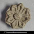 Fragment of wall-decoration made of dark grey clay with traces of red pigments and gilding (?) visible. The rosette-shaped ornament has a raised boss and eight pointed petals. The centre of each petal is incised with a deep line.