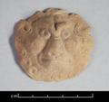 Round clay figure depicting a lion's face. The animal is shown in full frontal view with its head surrounded by the mane. Eyes, cheeks and forehead are bulging, giving the animal an evil expression. The fact that the plaque is flat on the reverse side suggests that it was applied to another object, probably a vessel.;