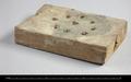 Oblong wooden block with nine drilled holes, possibly an incense-holder. Eight of the holes are arranged in a circle, the ninth is in the centre. Wedges inside the holes indicate that objects were inserted and kept upright. One side of the block shows clear traces of black paint.