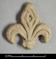 Floral ornament made of yellowish brown moulded clay with a light-coloured slip. Depicted is a fleur-de- lis. The leaves on both sides are decorated with rounded incised lines, while  the diamond-shaped leaf in the middle is decorated with diamond-shaped  incisions. At the lower part of the three leaves is a double-binding.