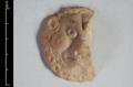 Fragment of a round clay plaque depicting a lion's face. The animal is shown in full frontal view with its head surrounded by a very stylised curled mane. Eyes, cheeks and forehead are bulging, giving the lion an evil expression. The fact that the plaque is flat on the reverse side suggests that it was applied to another object, probably a vessel.;