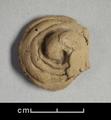 Fragment of figure made of reddish brown clay. Curled spiral that rises to point at centre. It represents a ringlet from the hair of the Buddha which, according to legend, grew back curly after he cut it as sign of his renunciation of earthly life.