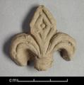 Floral ornament made of yellowish brown moulded clay, depicting a fleur-de-lis. The leaves on both sides are decorated with rounded incised lines, while the diamond-shaped leaf in the middle is decorated with diamond-shaped incisions. At the lower part of the three leaves is a double-binding.