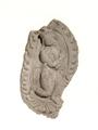 Stucco plaque showing a Gandharva kneeling in adoration on a lotus, both hands folded and facing to the right. Gandharvas are said to be supernatural beings with superb musical skills. This one wears a crown and a lot of jewellery and is surrounded by a flame-border.;