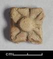 Fragment of a wall-decoration (?) made of brown clay with traces of red paint. The quatrefoil has a raised boss and angular petals each with an incised line in the middle.